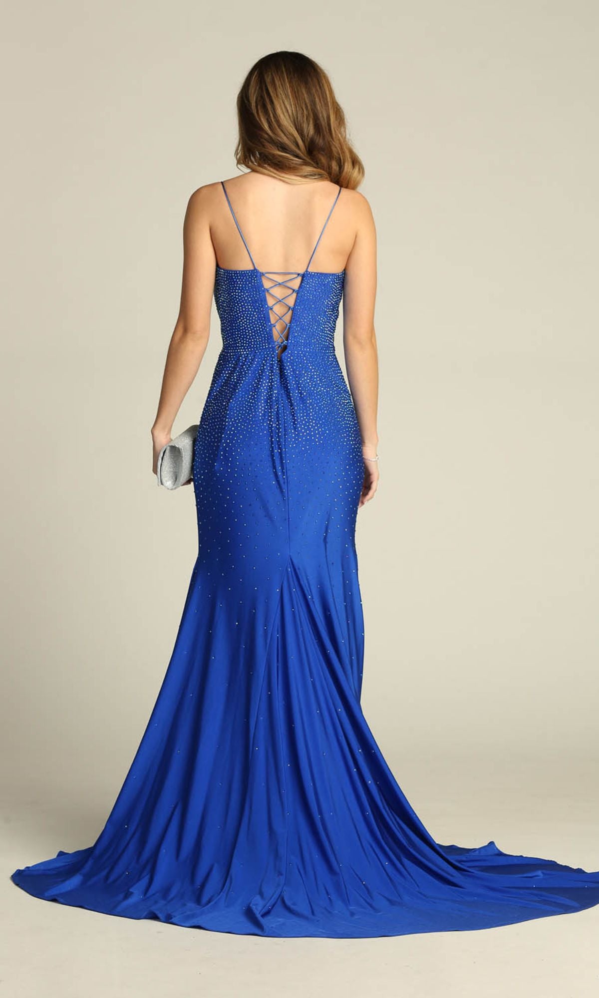 A woman in the Lace-Up Long Beaded Prom Dress: Chicas YG5026, a royal blue floor-length gown with thin straps and lace-up back, stands facing away holding a silver clutch.